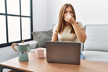 Sticker - Young brunette woman using laptop at home drinking a cup of coffee smelling something stinky and disgusting, intolerable smell, holding breath with fingers on nose. bad smell