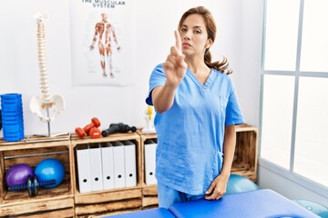 Wall Mural - Middle age hispanic physiotherapist woman working at pain recovery clinic pointing with finger up and angry expression, showing no gesture