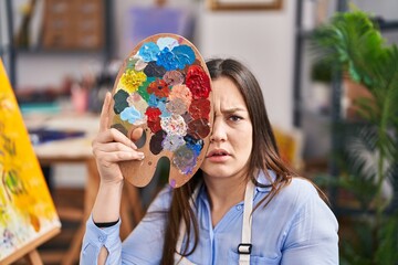 Wall Mural - Young brunette woman covering face with painter palette clueless and confused expression. doubt concept.