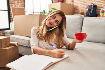 Canvas Print - Young blonde woman talking on the smartphone and writing on book at new home