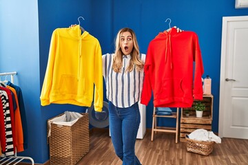 Poster - Young blonde woman holding clean laundry on hangers afraid and shocked with surprise and amazed expression, fear and excited face.