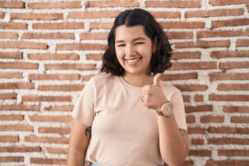 Young hispanic woman standing over bricks wall doing happy thumbs up gesture with hand. approving expression looking at the camera showing success.