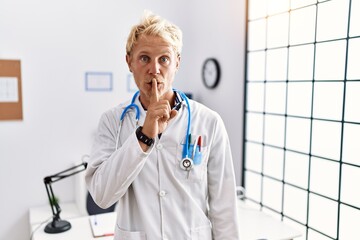 Poster - Young blond man wearing doctor uniform and stethoscope at clinic asking to be quiet with finger on lips. silence and secret concept.