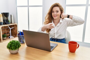 Poster - Young caucasian woman working at the office using computer laptop smiling doing talking on the telephone gesture and pointing to you. call me.