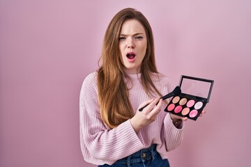 Wall Mural - Young caucasian woman holding makeup brush and blush palette angry and mad screaming frustrated and furious, shouting with anger. rage and aggressive concept.