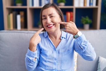 Sticker - Middle age hispanic woman sitting on the sofa at home smiling cheerful showing and pointing with fingers teeth and mouth. dental health concept.