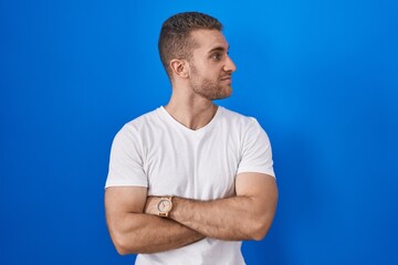 Sticker - Young caucasian man standing over blue background looking to the side with arms crossed convinced and confident