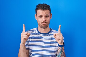 Young hispanic man standing over blue background pointing up looking sad and upset, indicating direction with fingers, unhappy and depressed.