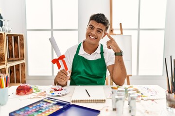 Sticker - Young hispanic man at art studio holding degree smiling pointing to head with one finger, great idea or thought, good memory