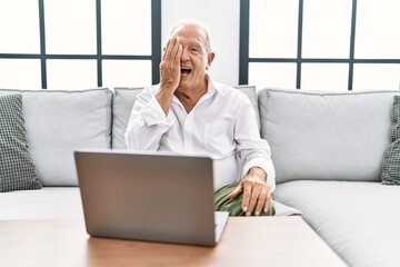 Sticker - Senior man using laptop at home sitting on the sofa covering one eye with hand, confident smile on face and surprise emotion.