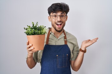 Poster - Arab man with beard holding green plant pot pointing aside with hands open palms showing copy space, presenting advertisement smiling excited happy