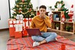 Arab young man using laptop sitting by christmas tree laughing and embarrassed giggle covering mouth with hands, gossip and scandal concept