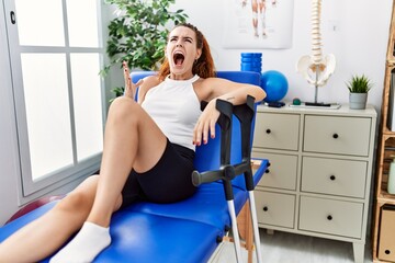 Poster - Young redhead woman lying on rehabilitation bed holding crutches crazy and mad shouting and yelling with aggressive expression and arms raised. frustration concept.