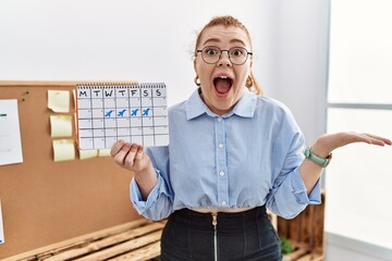 Poster - Young redhead woman holding travel calendar at the office celebrating victory with happy smile and winner expression with raised hands