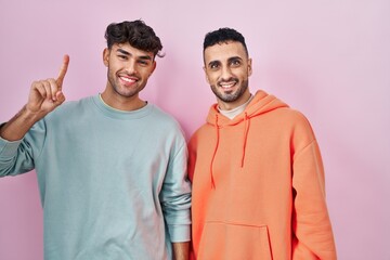 Wall Mural - Young hispanic gay couple standing over pink background showing and pointing up with finger number one while smiling confident and happy.