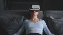 Woman In Virtual Reality Goggles Enters Metaverse Immersive Experience Via Headset Interface Resting On Sofa In Minimalistic Interior. Girl Gaming In Cyber Space. Futuristic Concept.