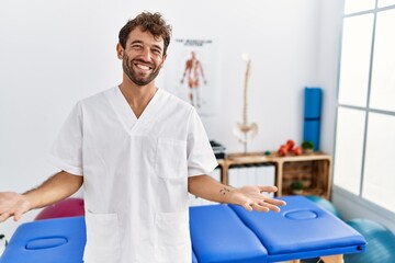 Canvas Print - Young handsome physiotherapist man working at pain recovery clinic smiling cheerful with open arms as friendly welcome, positive and confident greetings