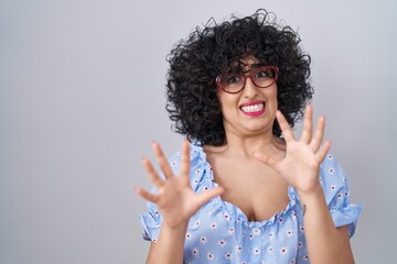 Poster - Young brunette woman with curly hair wearing glasses over isolated background afraid and terrified with fear expression stop gesture with hands, shouting in shock. panic concept.