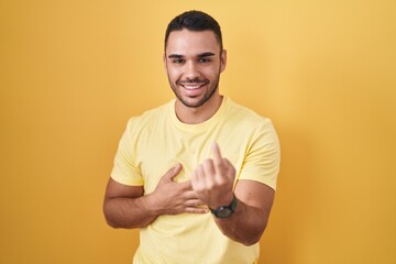 Poster - Young hispanic man standing over yellow background beckoning come here gesture with hand inviting welcoming happy and smiling