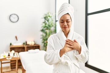 Wall Mural - Young brunette woman wearing towel and bathrobe standing at beauty center smiling with hands on chest with closed eyes and grateful gesture on face. health concept.