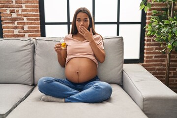 Canvas Print - Young pregnant woman holding moisturizer oil to hydrate belly covering mouth with hand, shocked and afraid for mistake. surprised expression