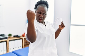 Poster - Young african woman holding covid record card at clinic annoyed and frustrated shouting with anger, yelling crazy with anger and hand raised