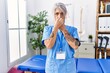 Middle age grey-haired woman wearing physiotherapist uniform at medical clinic smelling something stinky and disgusting, intolerable smell, holding breath with fingers on nose. bad smell