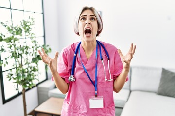 Poster - Young beautiful woman wearing doctor uniform and stethoscope crazy and mad shouting and yelling with aggressive expression and arms raised. frustration concept.