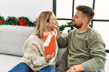 Wall Mural - Young caucasian couple smiling happy holding gift sitting on the sofa by christmas tree at home.