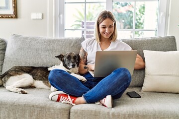 Poster - Young caucasian girl smiling happy sitting on the sofa with dog using laptop at home.