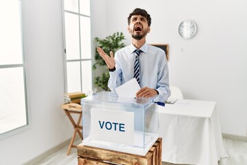 Wall Mural - Hispanic man with beard voting putting envelop in ballot box crazy and mad shouting and yelling with aggressive expression and arms raised. frustration concept.