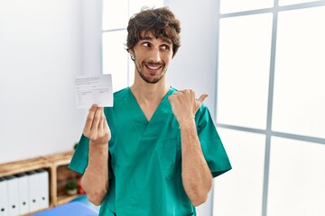 Sticker - Young hispanic doctor man holding covid record card smiling with happy face looking and pointing to the side with thumb up.