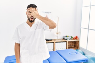 Sticker - Young handsome man with beard working at pain recovery clinic peeking in shock covering face and eyes with hand, looking through fingers with embarrassed expression.