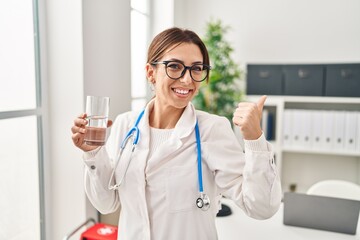 Sticker - young brunette doctor woman holding glass of water smiling happy pointing with hand and finger to th