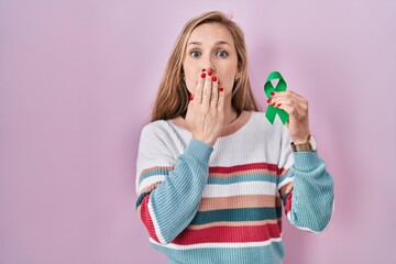 Wall Mural - Young blonde woman holding support green ribbon covering mouth with hand, shocked and afraid for mistake. surprised expression