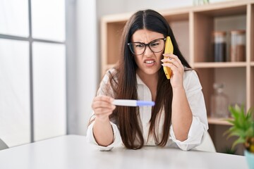 Poster - Young brunette woman holding pregnancy test result speaking on the phone skeptic and nervous, frowning upset because of problem. negative person.