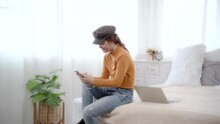 Beautiful asian woman sitting on bed using smartphone chatting with boyfriend with happy, Female wearing hat use free time rest from work on laptop by play iphone in bedroom.
