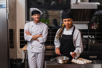 young african american sous chef in apron holding egg near bowl and whisk while cooking near chef in kitchen.