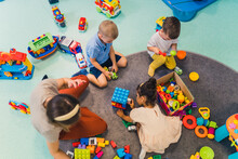 Toddlers And Their Nursery Teacher Playing With Plastic Building Blocks And Colorful Car Toys While Sitting On The Floor In A Playroom. Early Brain And Skills Development. High Quality Photo