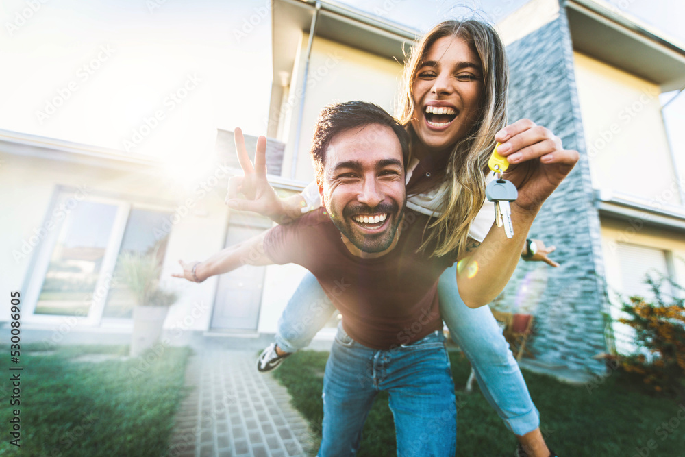 Happy young couple holding home keys after buying real estate - Husband and wife standing outside in front of their new house