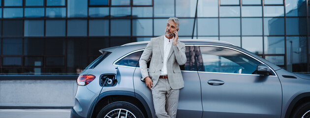 Businessman holding smartphone while charging car at electric vehicle charging station, closeup.