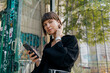 © PhotoBook - Elegant attractive girl in black shirt listens music in wireless headphones and posing at camera. Charming stylish lady spend time outdoor in city park and scrolling smartphone