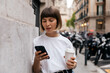 © PhotoBook - Cute young caucasian girl smiling looking at smartphone relaxing walking in city. Short-haired woman wears casual clothes, holds coffee and smartphone. Concept of enjoying moment