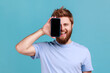 © khosrork - Portrait of smiling optimistic bearded man covering half face with cellphone and looking at camera with toothy smile, positive glad face. Indoor studio shot isolated on blue background.