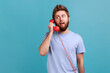 © khosrork - Portrait of handsome young adult bearded man in T-shirt talking retro phone handset to camera, looking away, having conversation. Indoor studio shot isolated on blue background.