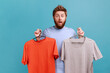 © khosrork - Portrait of surprised astonished handsome bearded man holding two hangers with gray and orange T-shirts, expressing shock, keeps mouth opened. Indoor studio shot isolated on blue background.
