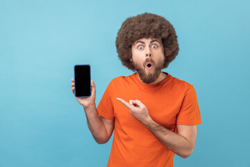 Wall Mural - portrait of shocked satisfied man with afro hairstyle wearing orange t-shirt pointing at mobile phon