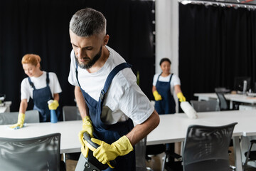 Wall Mural - man with floor scrubber machine near interracial women cleaning desks on blurred background.