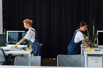 Wall Mural - side view of multiethnic women cleaning office near computer monitors with blank screen.