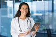 © Liubomir - Portrait of young beautiful female doctor with tablet computer, Hispanic female doctor working in modern clinic office smiling and looking at camera wearing white medical coat and stethoscope.